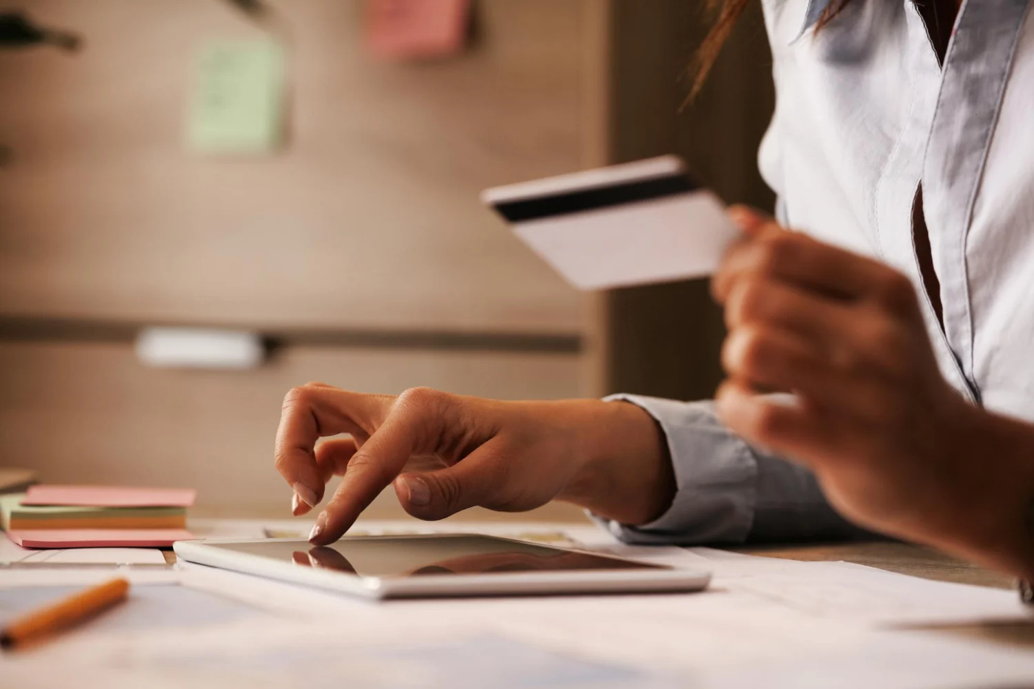 A hand holding a payment card while tapping a tablet on a desk