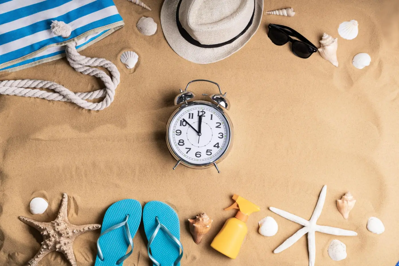 An alarm clock on the sand among a beach hat, sunglasses, flip-flops and shells